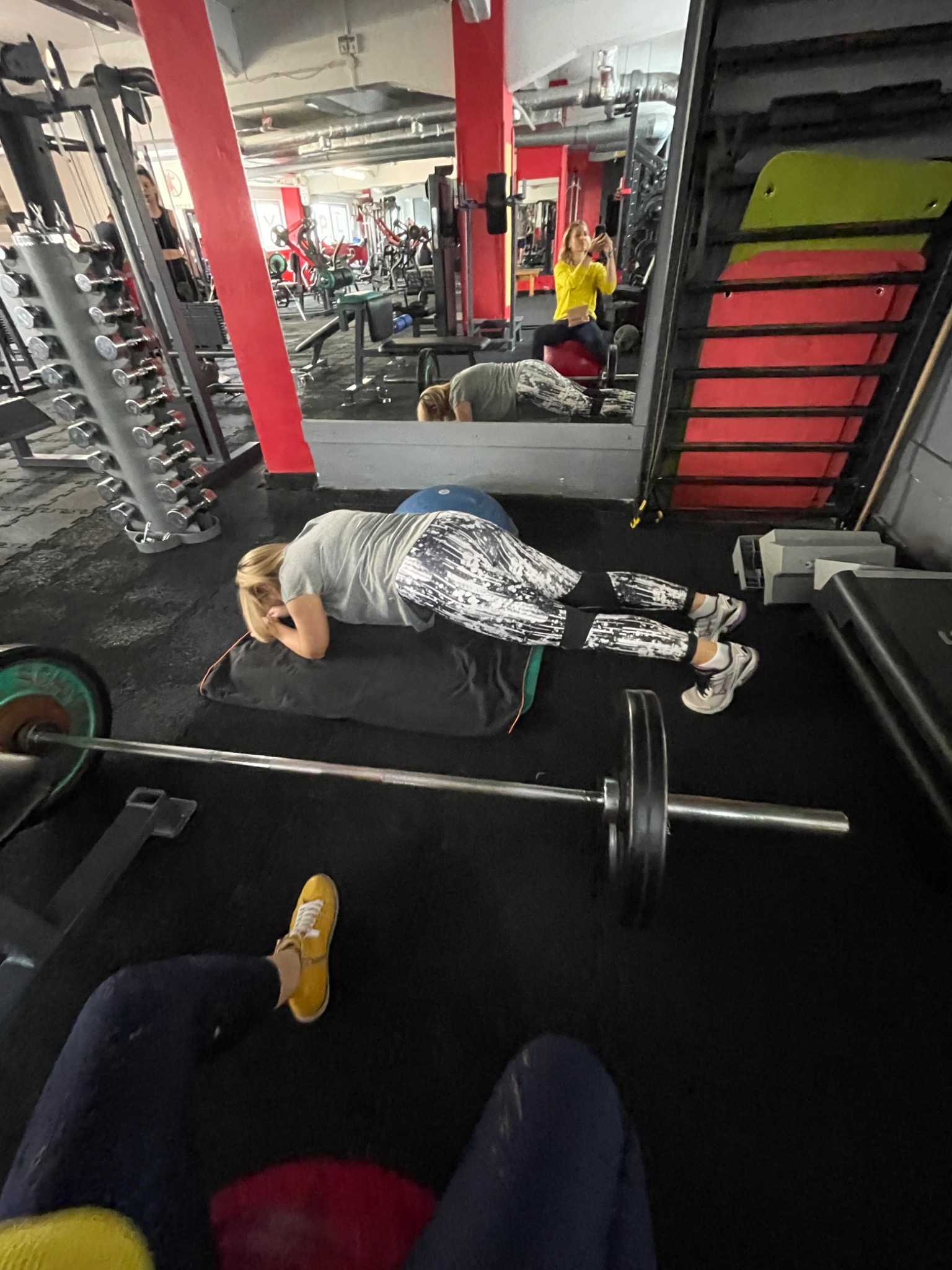 Kobieta w Sines wykonuje ćwiczenie na macie z obciążeniem na plecach w siłowni, widoczne odbicie w lustrze i inne osoby ćwiczące.