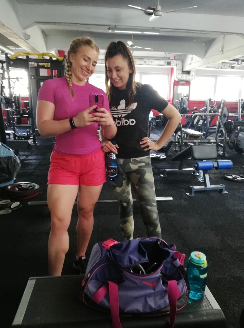 Dwie uśmiechnięte kobiety w siłowni robią selfie; jedna w różowej koszulce i spodenkach, druga w czarnej koszulce Adidas i legginsach moro; torba sportowa Nike i butelka wody na pierwszym planie.