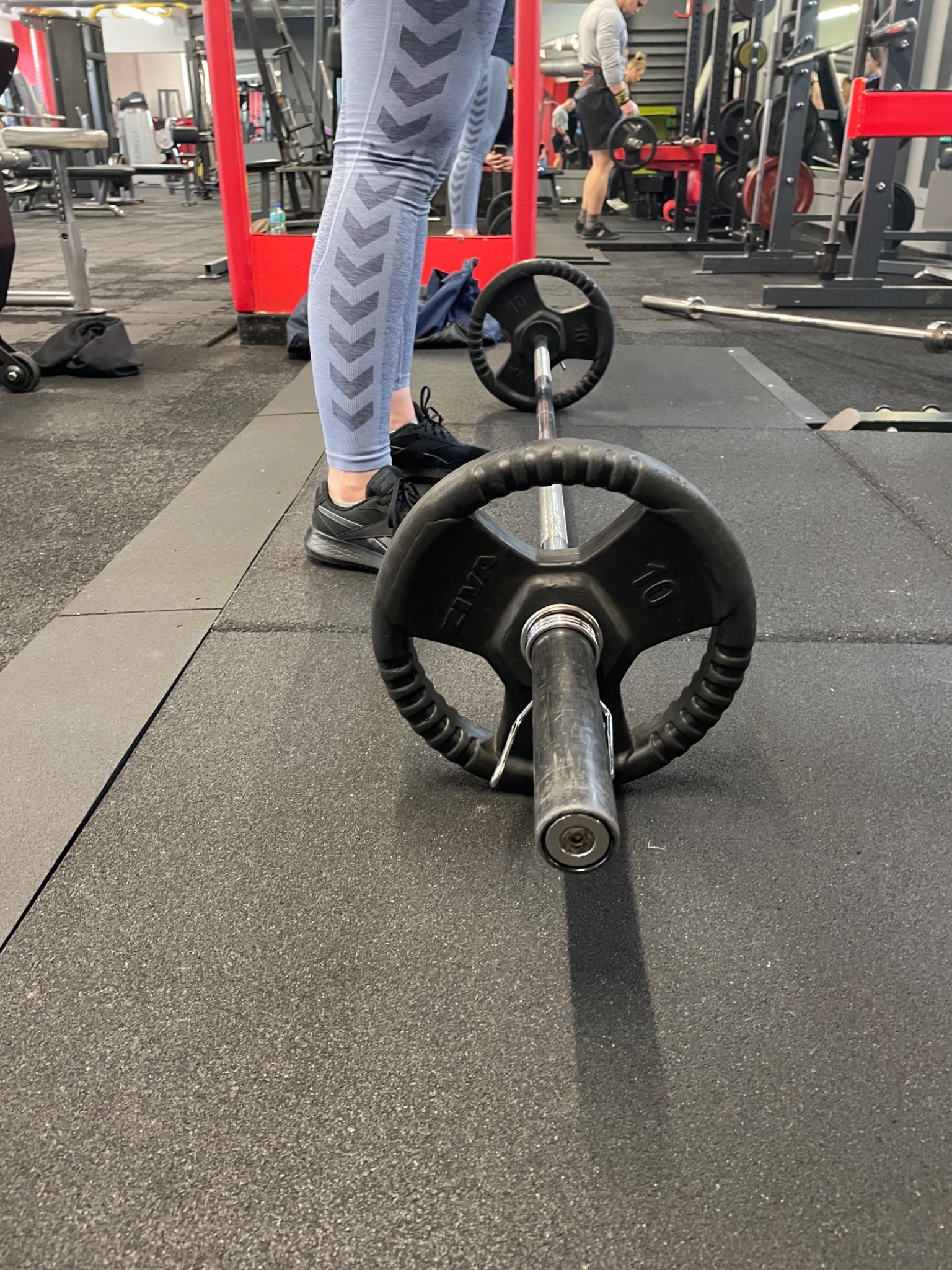 Ciężar 10 kg na gryfie w siłowni, widok z poziomu podłogi, osoba w legginsach z wzorem w tle.