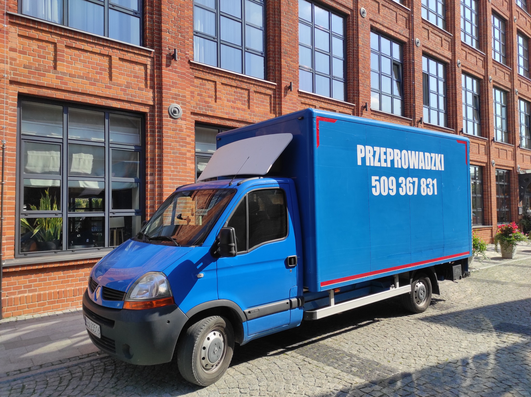 Niebieski samochód dostawczy z napisem 'Przeprowadzki 509 367 831' na tle ceglanego budynku z dużymi oknami.