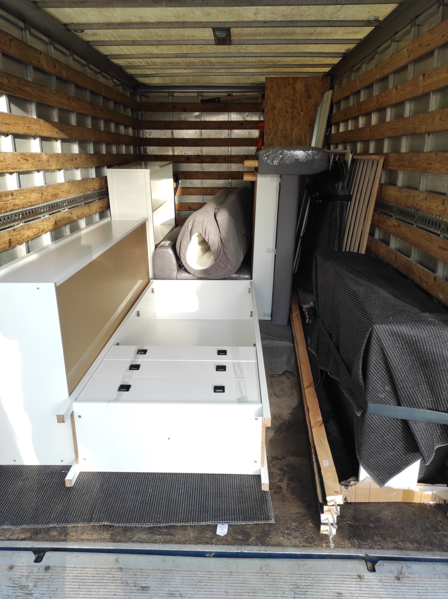 Wnętrze busa załadowane meblami podczas przeprowadzki: biała komoda z szufladami, zwinięty materac i elementy łóżka zabezpieczone folią, widoczne drewniane ściany i podłoga pojazdu.