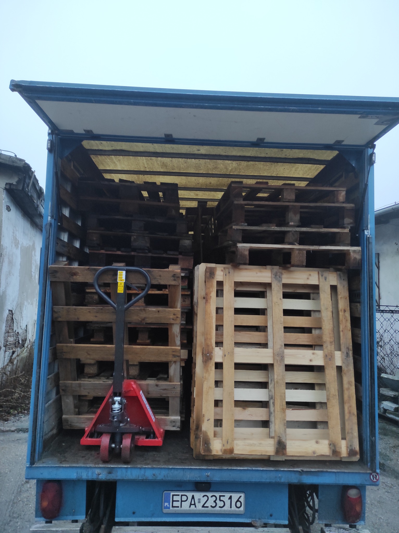 Wnętrze otwartego busa załadowane drewnianymi paletami i czerwonym wózkiem paletowym, widok z tyłu pojazdu.