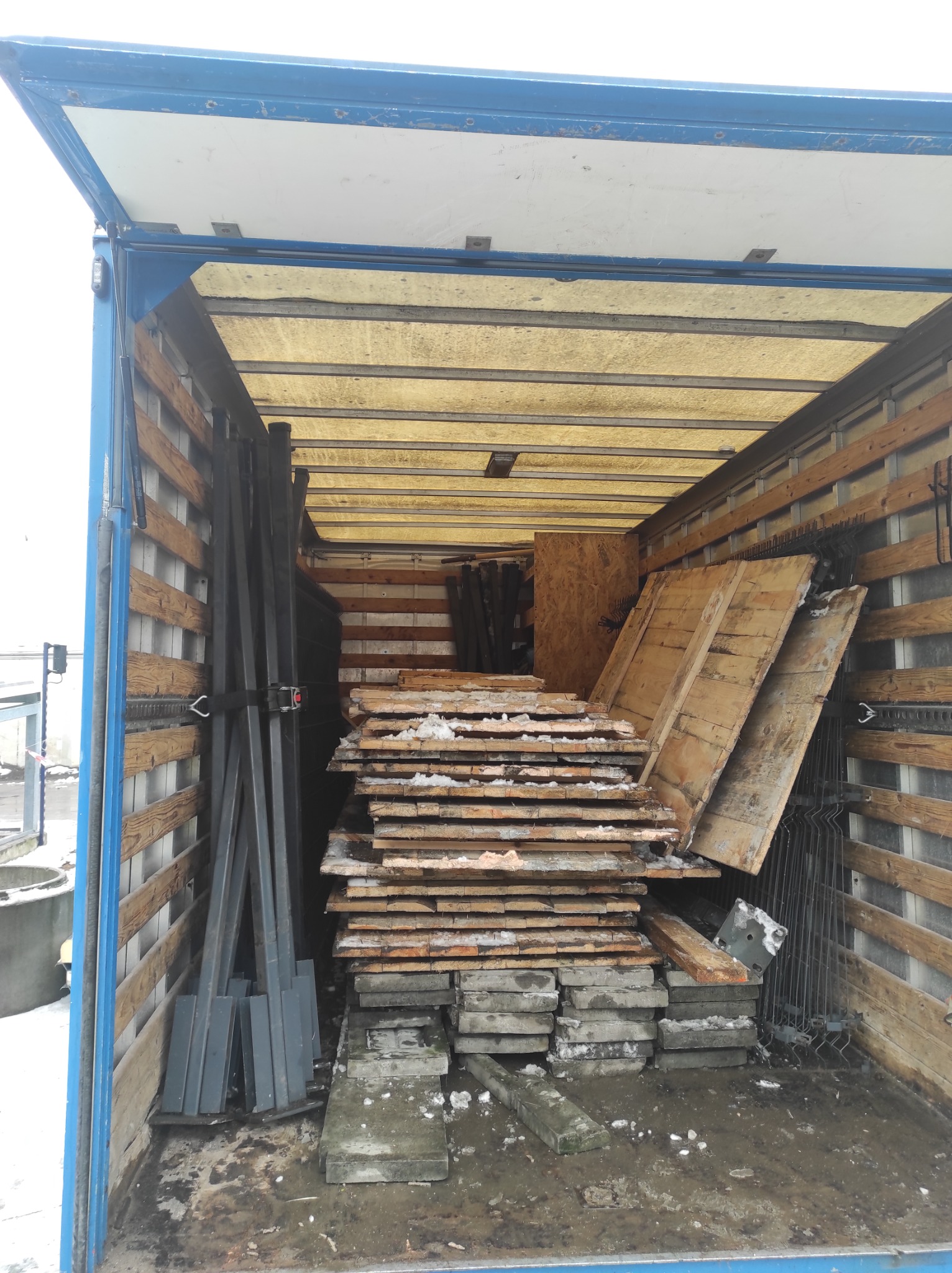 Wnętrze otwartego busa załadowane materiałami budowlanymi: ułożone deski z resztkami śniegu, betonowe płyty chodnikowe i metalowe profile zabezpieczone pasem transportowym.