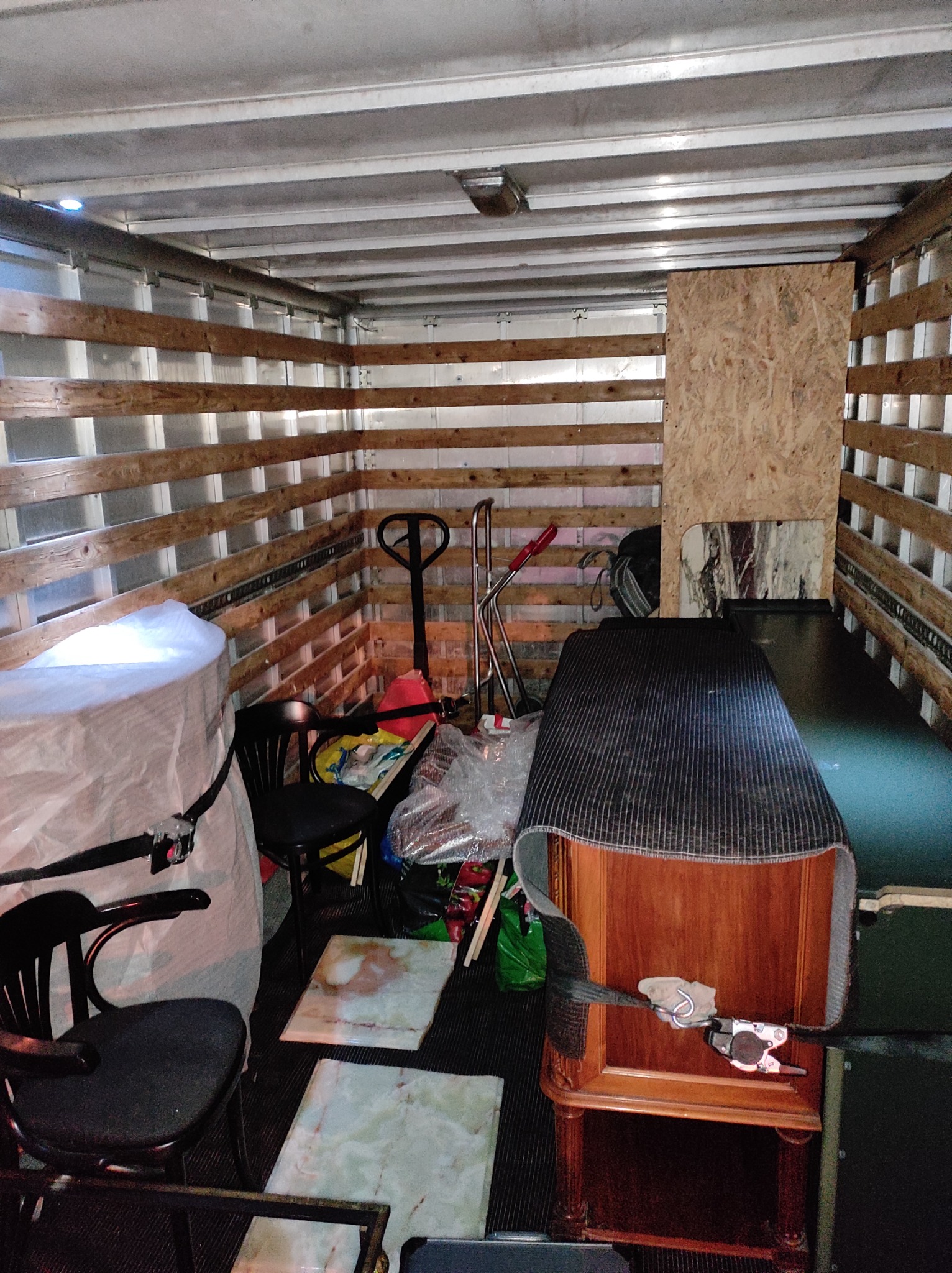 Wnętrze przestrzeni ładunkowej pojazdu z zabezpieczonymi meblami i przedmiotami, w tym krzesłami, biurkiem i paleciakiem, przygotowanymi do transportu.