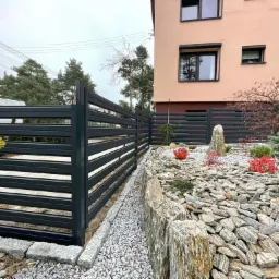 Nowoczesne ogrodzenie z poziomych, metalowych profili w kolorze antracytowym, otoczone ozdobnym ogrodem skalnym z kamieniami i roślinami, na tle budynku mieszkalnego.