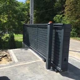 Grafitowa brama przesuwna z poziomymi panelami, z pomarańczowym światłem ostrzegawczym, zamontowana na szarej kostce brukowej z jasną obwódką, na tle zieleni i słupa energetycznego.