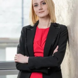 Portret kobiety w czarnym żakiecie i czerwonej bluzce, opierającej się o ścianę z dekoracyjnym tynkiem w biurze z oknem w tle.