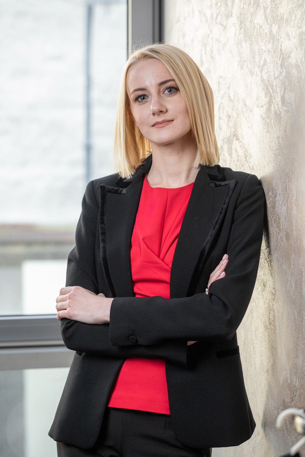 Portret kobiety w czarnym żakiecie i czerwonej bluzce, opierającej się o ścianę z dekoracyjnym tynkiem w biurze z oknem w tle.