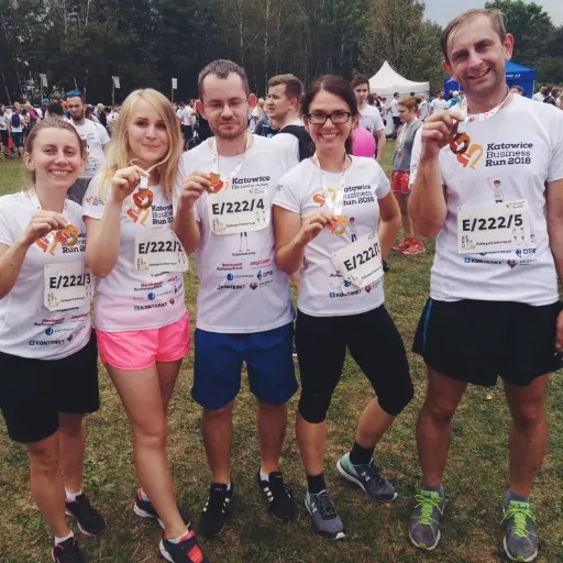 Grupa pięciu osób w białych koszulkach Katowice Business Run 2018, z medalami na szyjach, stojąca na trawie, z widocznymi logotypami sponsorów na koszulkach, w tle widoczny tłum uczestników.
