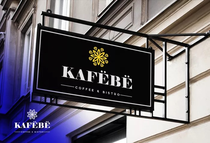 Znak reklamowy kawiarni 'Kafebë' z czarnym tłem, białym napisem i żółtą ozdobną rozetą, zamontowany na metalowej konstrukcji na tle jasnej elewacji budynku.