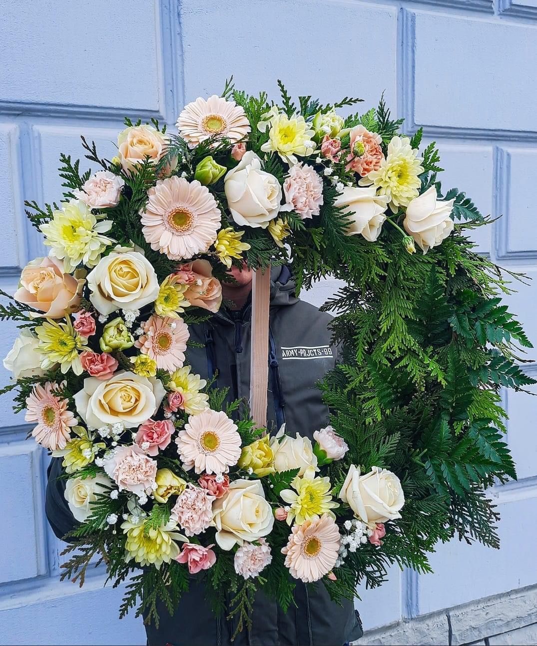 Okrągły wieniec z białych i pastelowych róż, gerber i goździków, oparty na zielonych gałązkach, trzymany przez osobę w kurtce na tle jasnoniebieskiej ściany.