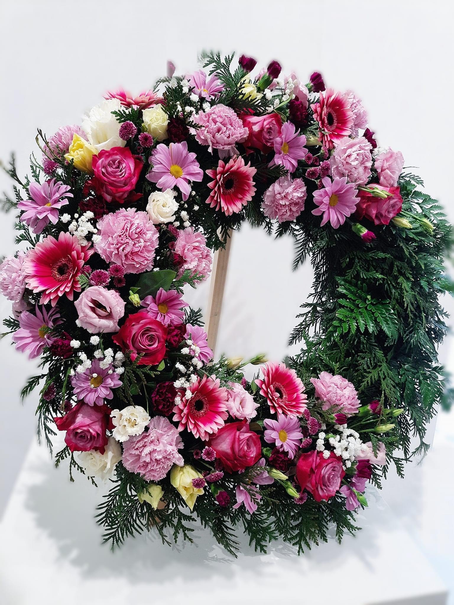 Wieniec pogrzebowy z różowych i białych róż, gerber, goździków i drobnych białych kwiatów na zielonym podkładzie z igliwia, ustawiony na drewnianym stojaku.