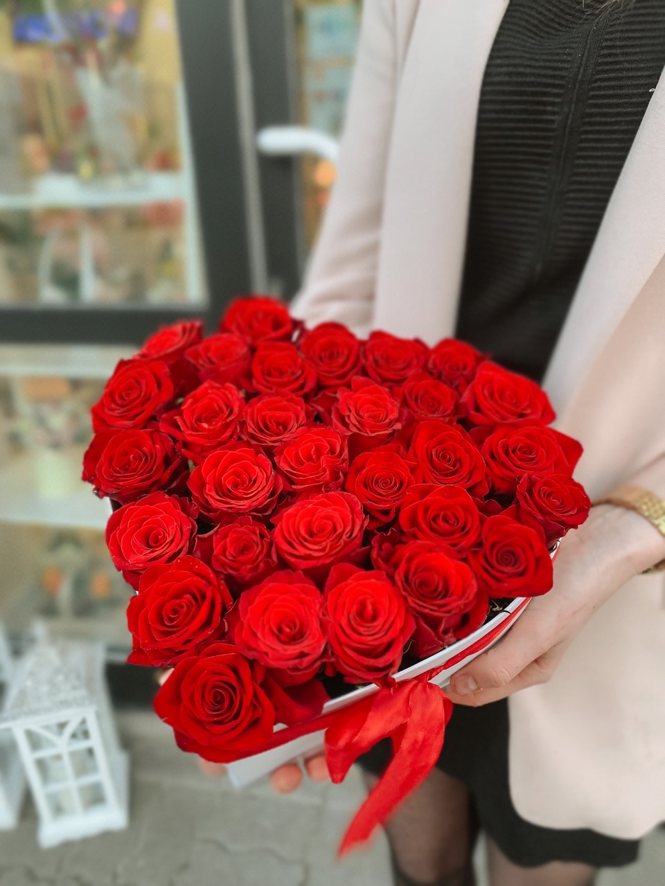 Osoba trzyma w dłoniach białe pudełko w kształcie serca wypełnione czerwonymi różami, przewiązane czerwoną wstążką. W tle widoczne rozmyte witryny sklepowe.