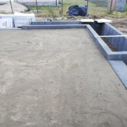 Przygotowany teren pod przyszły taras z obrzeżami z szarej kostki, widoczne materiały budowlane i worki na śmieci w tle.