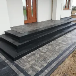 Nowoczesne schody wejściowe z czarnych i szarych kostek brukowych prowadzące do domu z drewnianymi drzwiami i oknem.