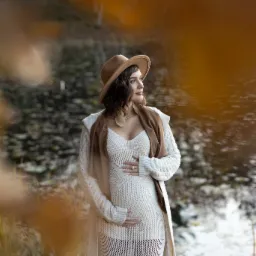 Kobieta w ciąży w beżowej sukience, kapeluszu i kardiganie pozuje na tle jesiennego krajobrazu z jeziorem, trzymając ręce na brzuchu i spoglądając w bok.