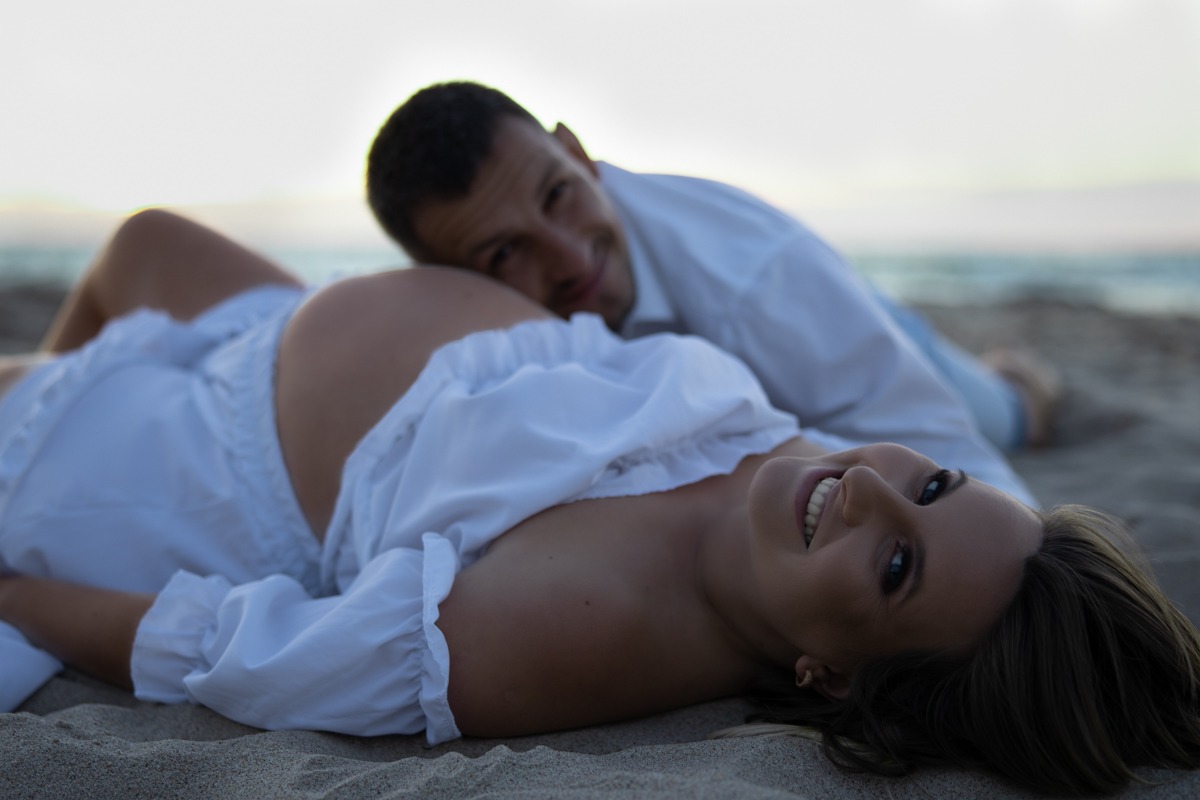Uśmiechnięta kobieta w białej bluzce z falbankami, leżąca na piasku z widocznym ciążowym brzuchem, obok niej uśmiechnięty mężczyzna w białej koszuli, w tle morze.