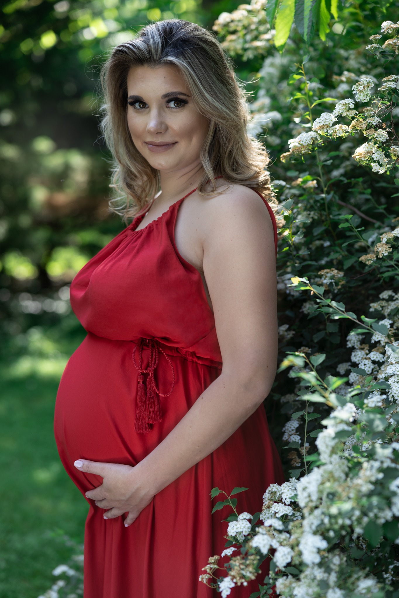 Kobieta w czerwonej sukience ciążowej pozuje na tle kwitnących krzewów w Białogardzie, delikatny uśmiech, dłoń na brzuchu.