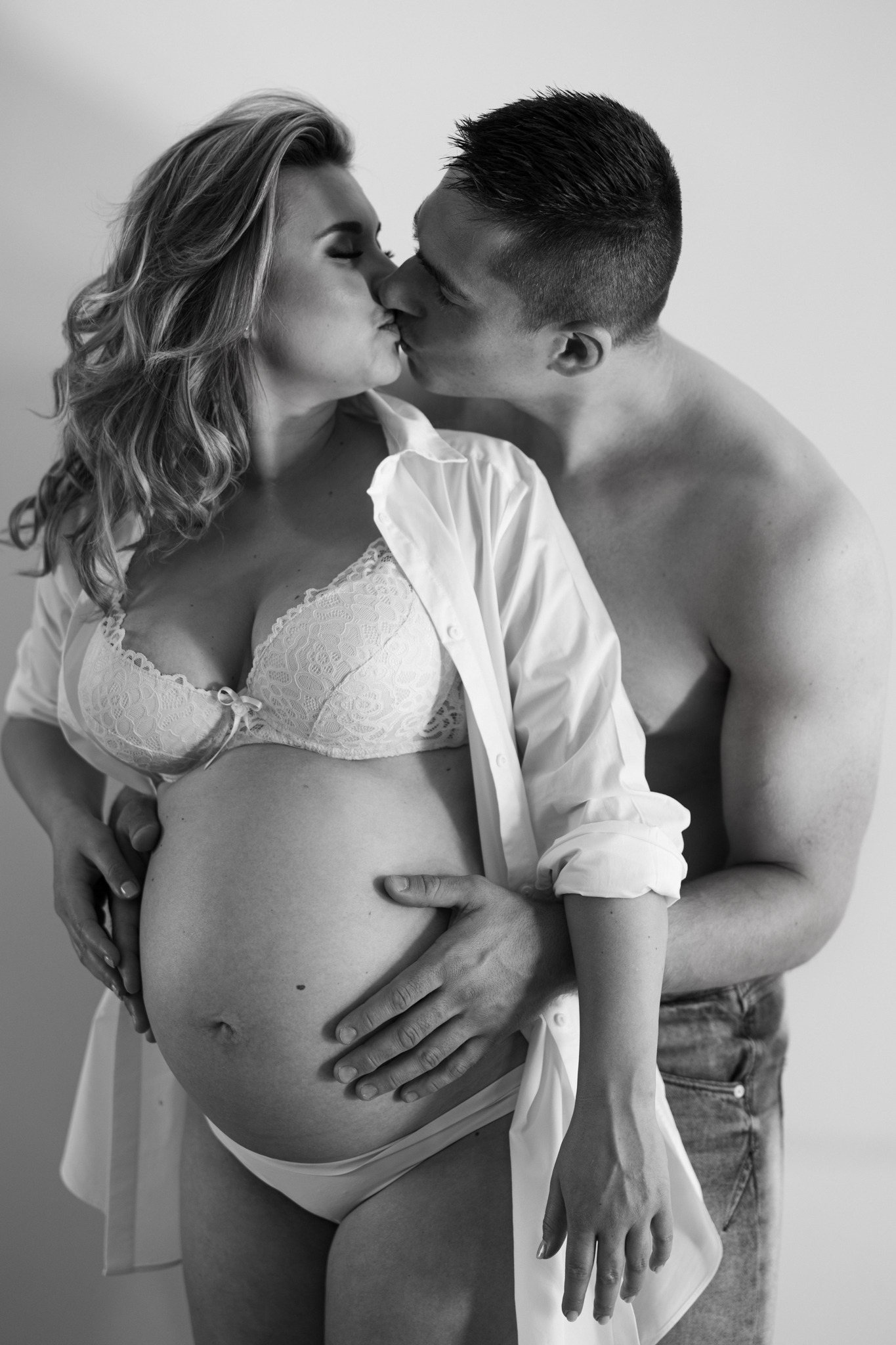 Czarno-białe zbliżenie na całującą się parę, gdzie kobieta w bieliźnie i rozpiętej koszuli eksponuje ciążowy brzuszek, a mężczyzna obejmuje ją od tyłu.