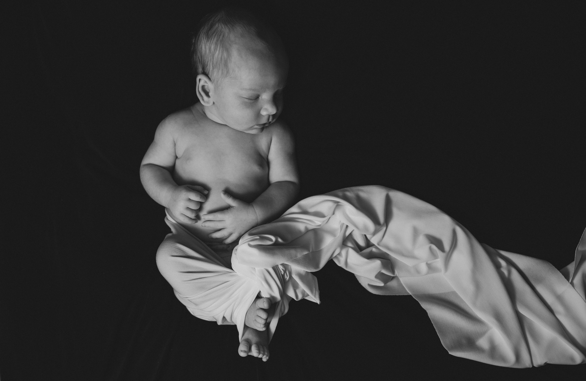 Monochromatyczne ujęcie śpiącego noworodka owiniętego w delikatną, białą tkaninę na ciemnym tle, z dłońmi złożonymi na brzuszku.