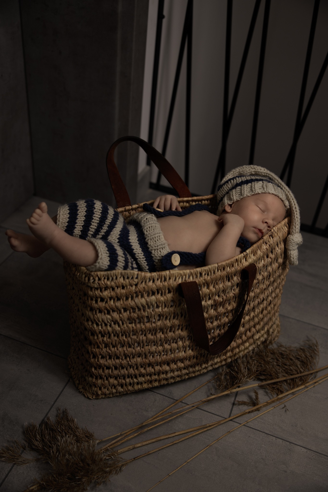 Noworodek śpiący w wiklinowym koszu w pasiastym, dzierganym stroju i czapce z pomponem, obok leżą źdźbła trawy.