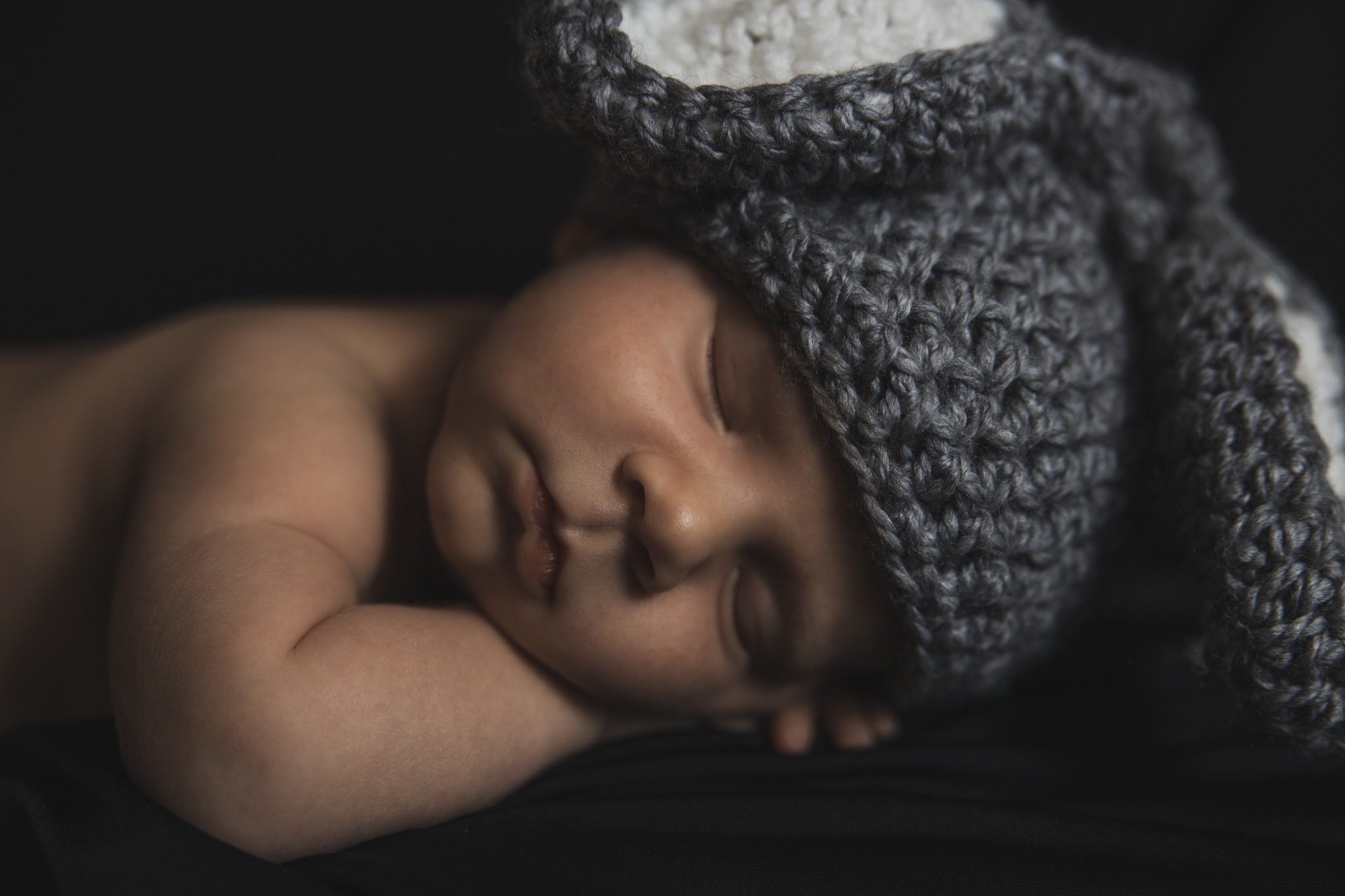 Śpiący noworodek w szarej, dzierganej czapce z uszami królika na ciemnym tle.