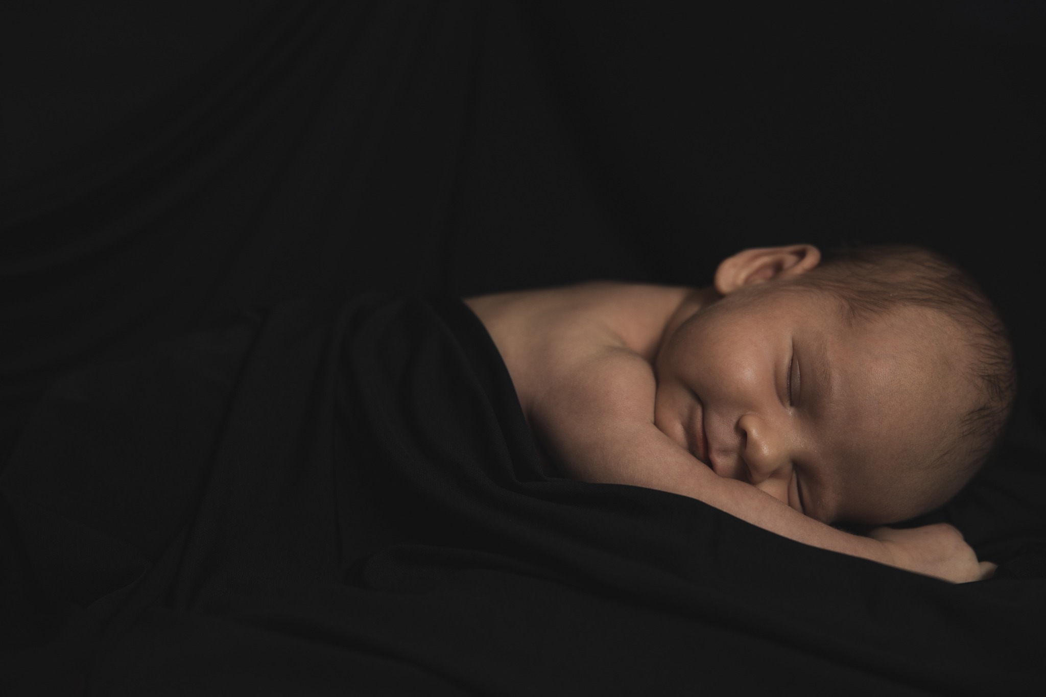 Śpiący noworodek otulony czarną tkaniną, delikatne oświetlenie podkreśla rysy twarzy.
