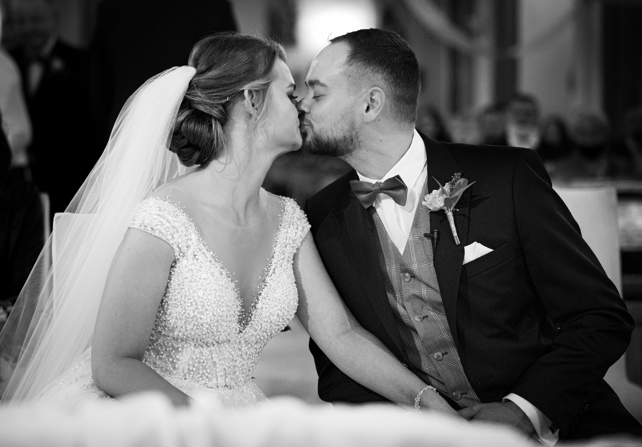 Czarno-białe zbliżenie całującej się pary młodej: panna młoda w bogato zdobionej sukni i welonie, pan młody w garniturze z muszką i butonierką.