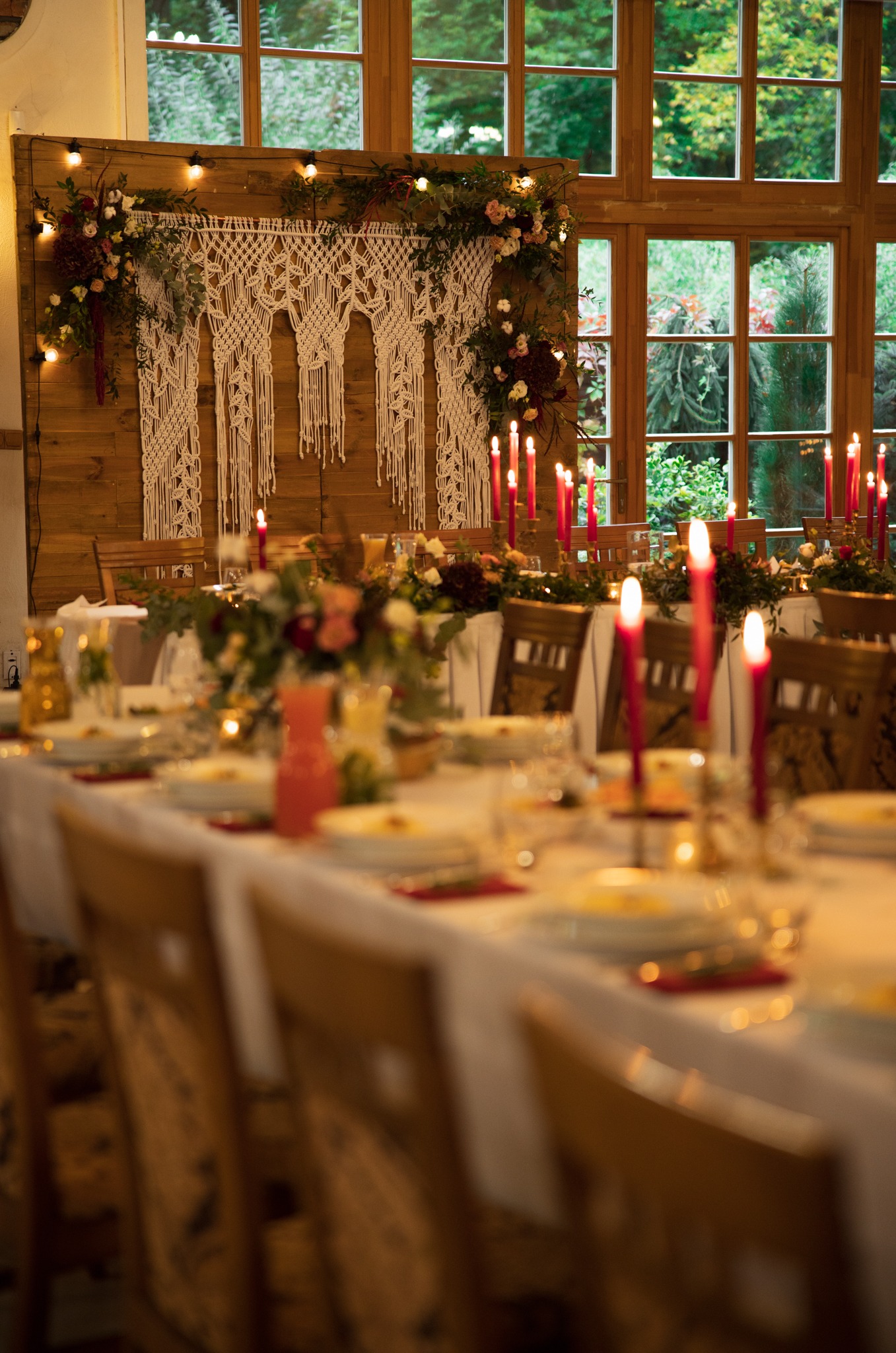 Uroczysta aranżacja stołu z płonącymi czerwonymi świecami, kwiatami i makramą na drewnianej ścianie w tle, widok na zielony ogród przez okno.