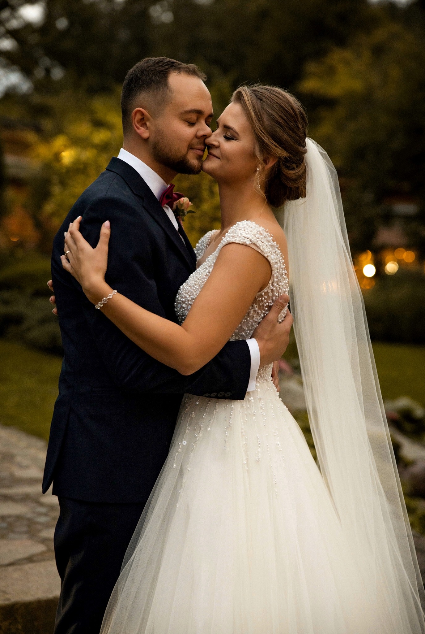Młoda para w objęciach, panna młoda w bogato zdobionej sukni ślubnej i welonie, pan młody w granatowym garniturze z bordową muchą, delikatne oświetlenie, parkowe tło.