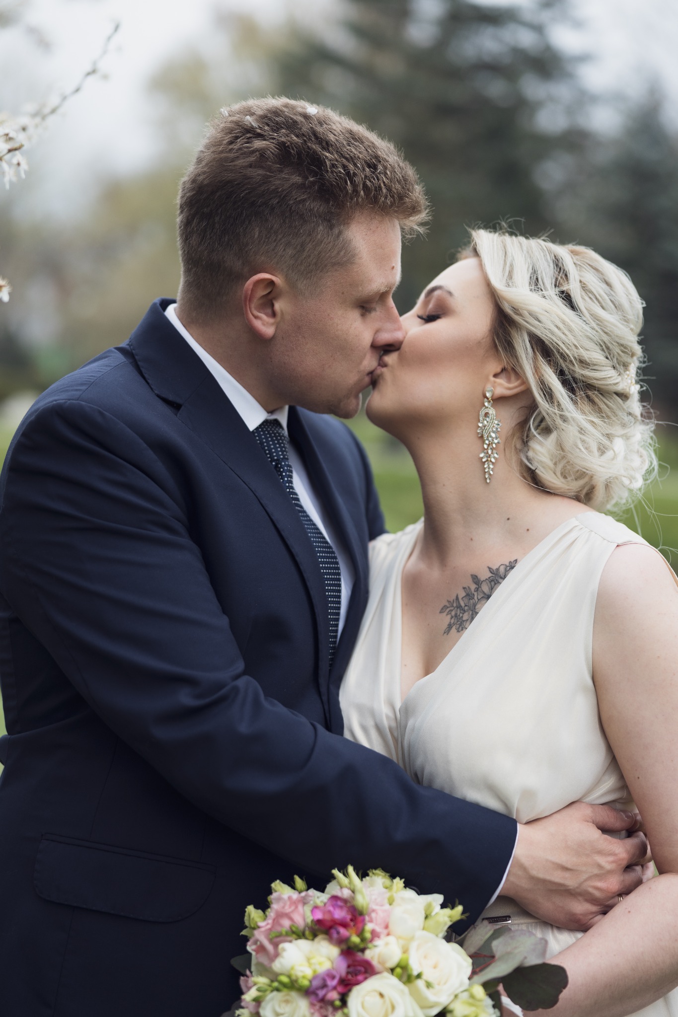 Pocałunek pary młodej w plenerze, panna młoda z tatuażem na dekolcie i długimi kolczykami, pan młody w granatowym garniturze, bukiet ślubny w dłoniach.