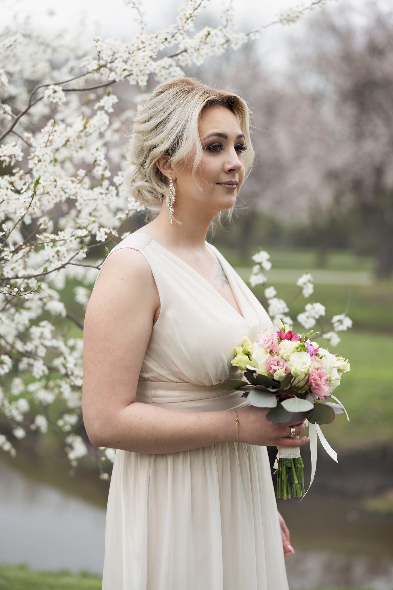 Kobieta w jasnej sukni trzyma bukiet kwiatów na tle kwitnącego drzewa, z widoczną rzeką w tle.