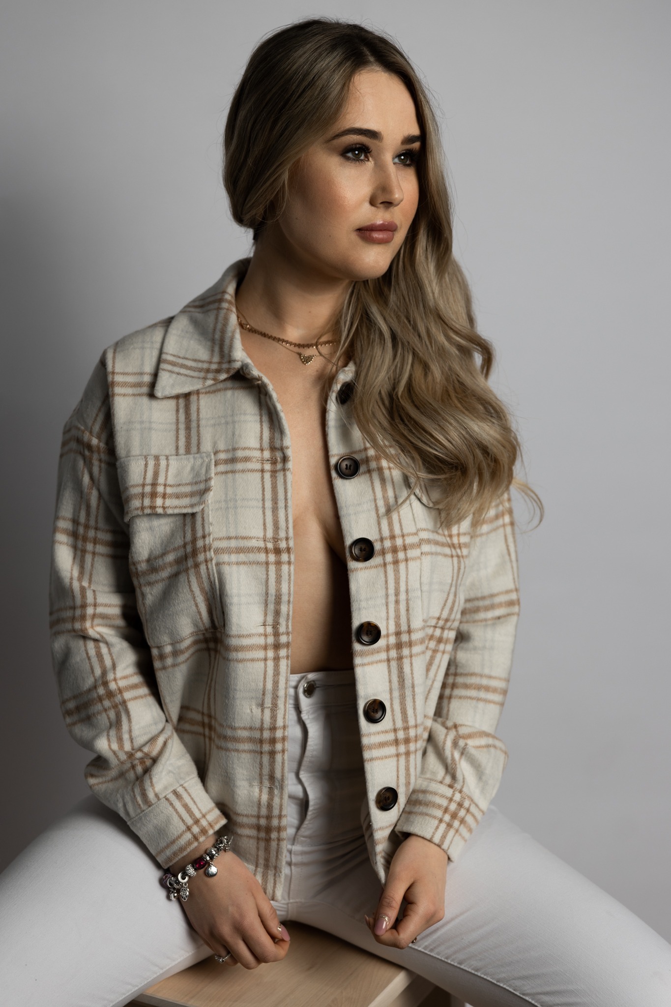 Portret kobiety z blond włosami, w rozpiętej koszuli w kratę i białych spodniach, siedzącej na stołku na jasnym tle.