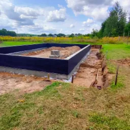 Wylany fundament z izolacją pionową w wykopie, widoczne szalunki i paleta drewniana na trawie, w tle łąka i las.