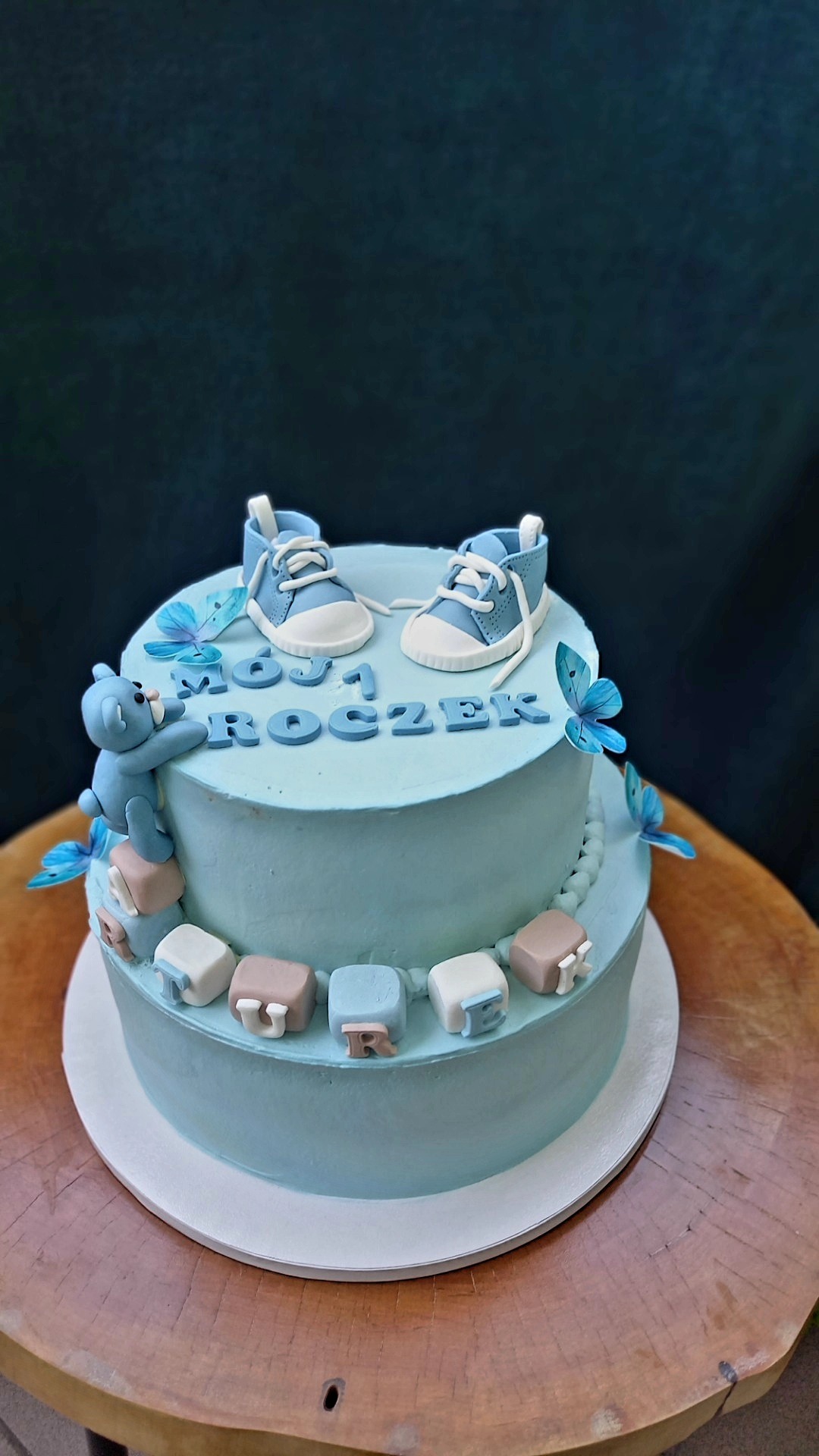 Dwupoziomowy tort w odcieniach błękitu z napisem 'Mój 1 Roczek', ozdobiony figurkami butów niemowlęcych, misia i motyli, umieszczony na drewnianym stole.