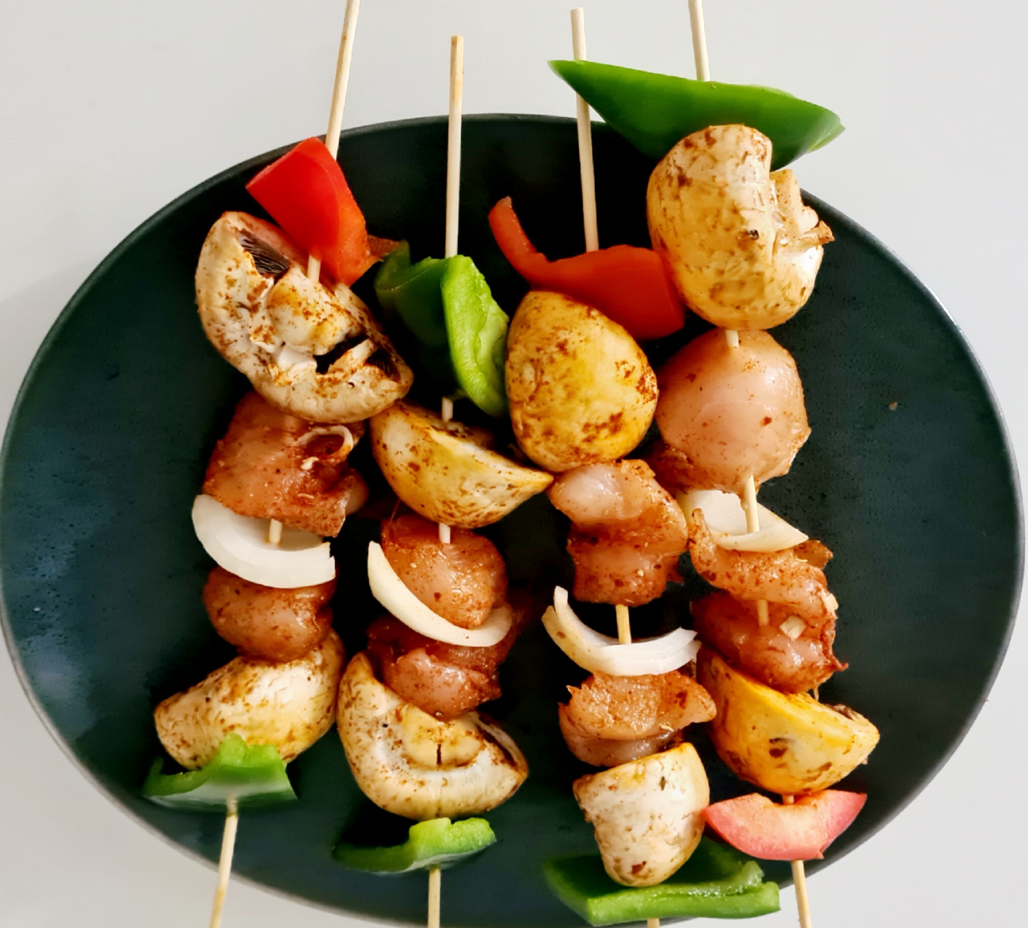 Trzy szaszłyki z kurczaka, pieczarek, cebuli i papryki na ciemnym talerzu, widok z góry na jasnym tle.