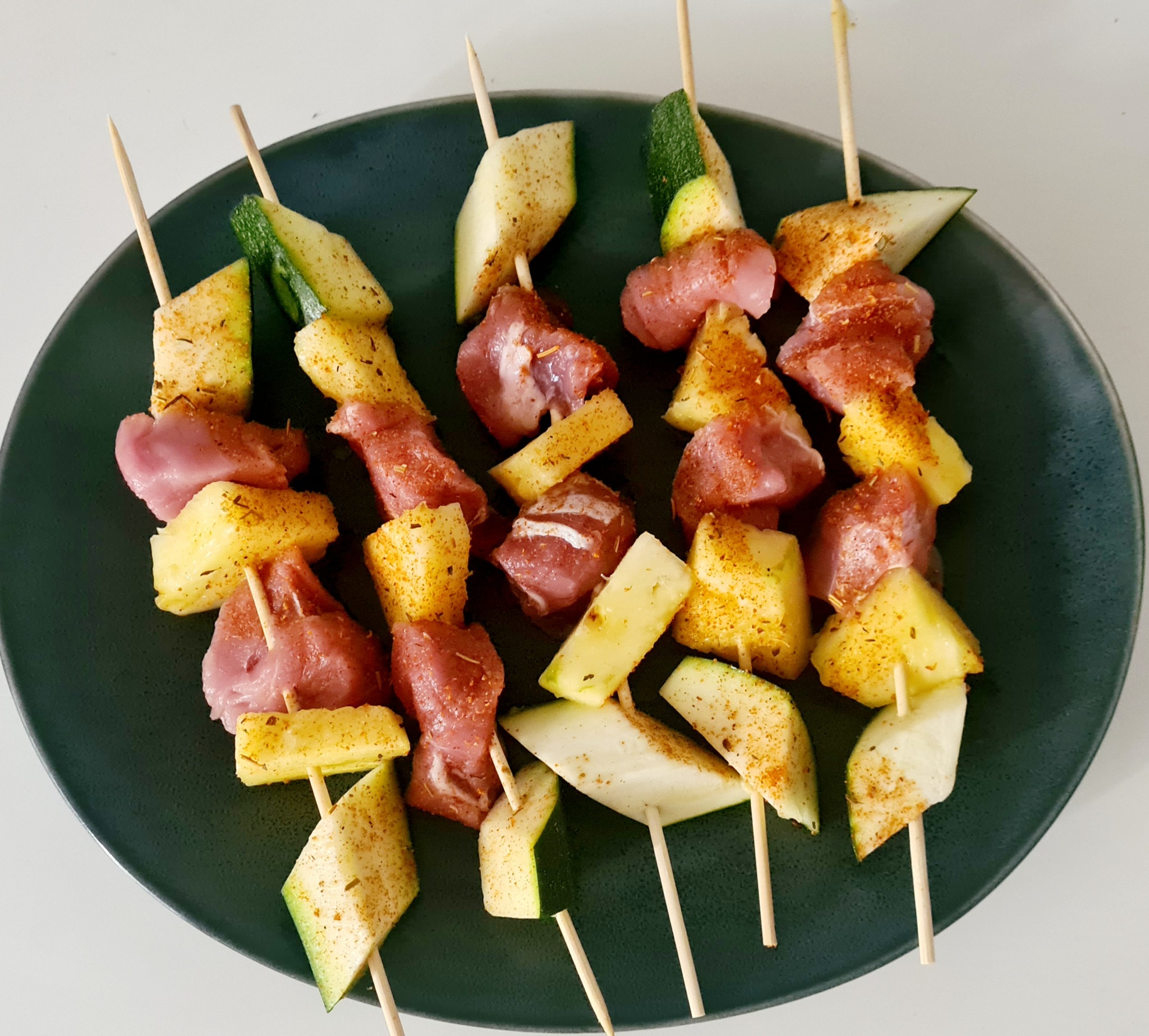 Szklana płyta z czterema szaszłykami z surowego mięsa, ananasa i cukinii, przyprawione, gotowe do pieczenia.