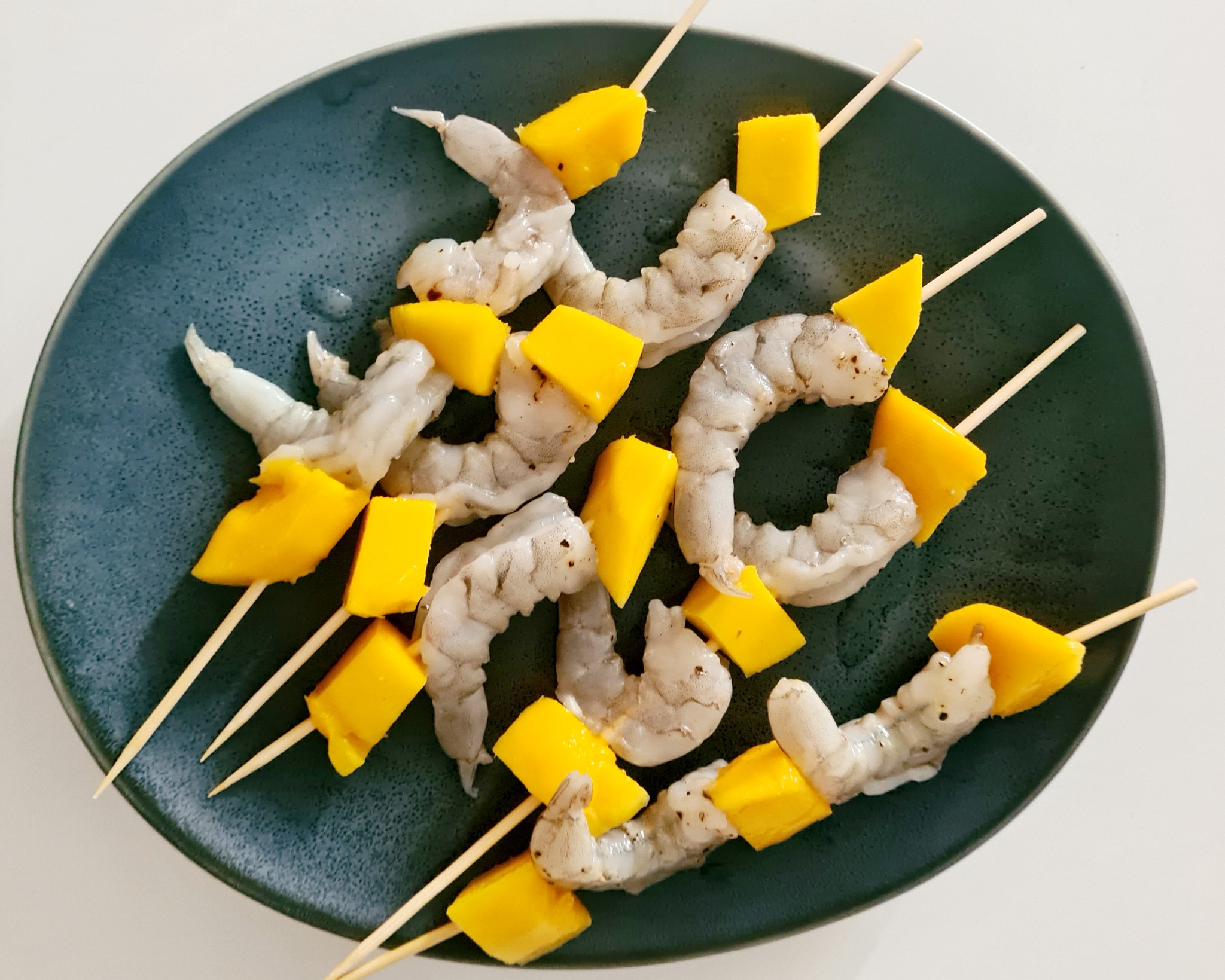 Szpadki z surowych krewetek i kostek mango na ciemnym, nakrapianym talerzu, przygotowane do grillowania lub smażenia.