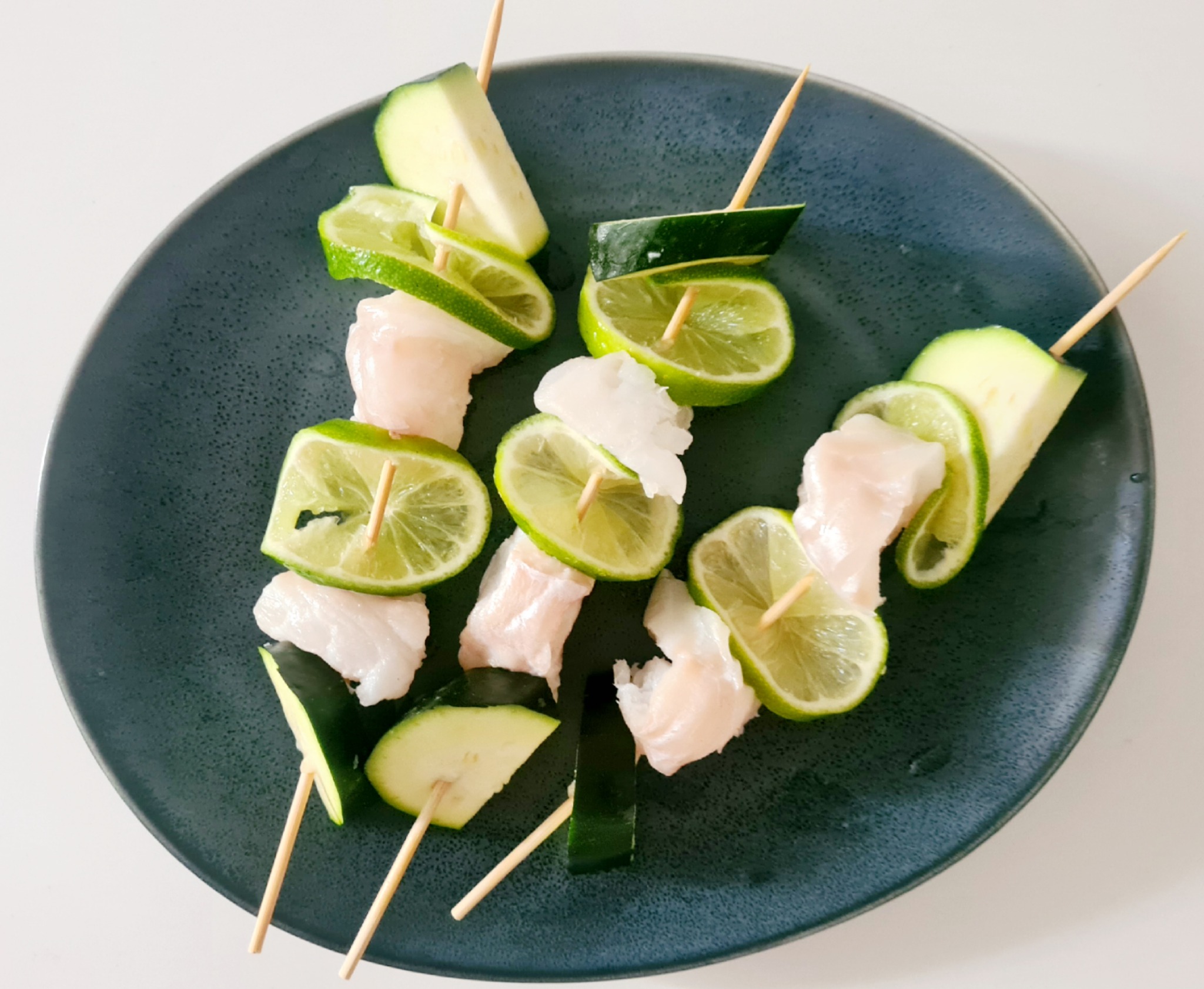 Trzy szaszłyki z białej ryby, limonki i cukinii, ułożone na ciemnym, nakrapianym talerzu na białym tle.