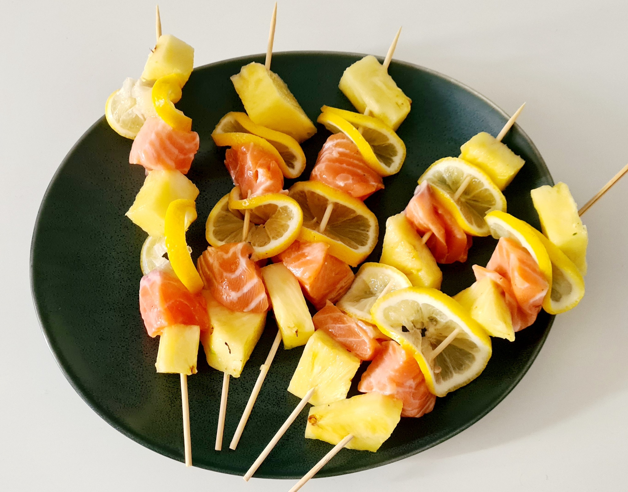 Szklana taca z ciemną obwódką prezentuje kolorowe szaszłyki z ananasa, łososia i cytryny na drewnianych patyczkach, ułożone w artystycznym nieładzie na białym tle.
