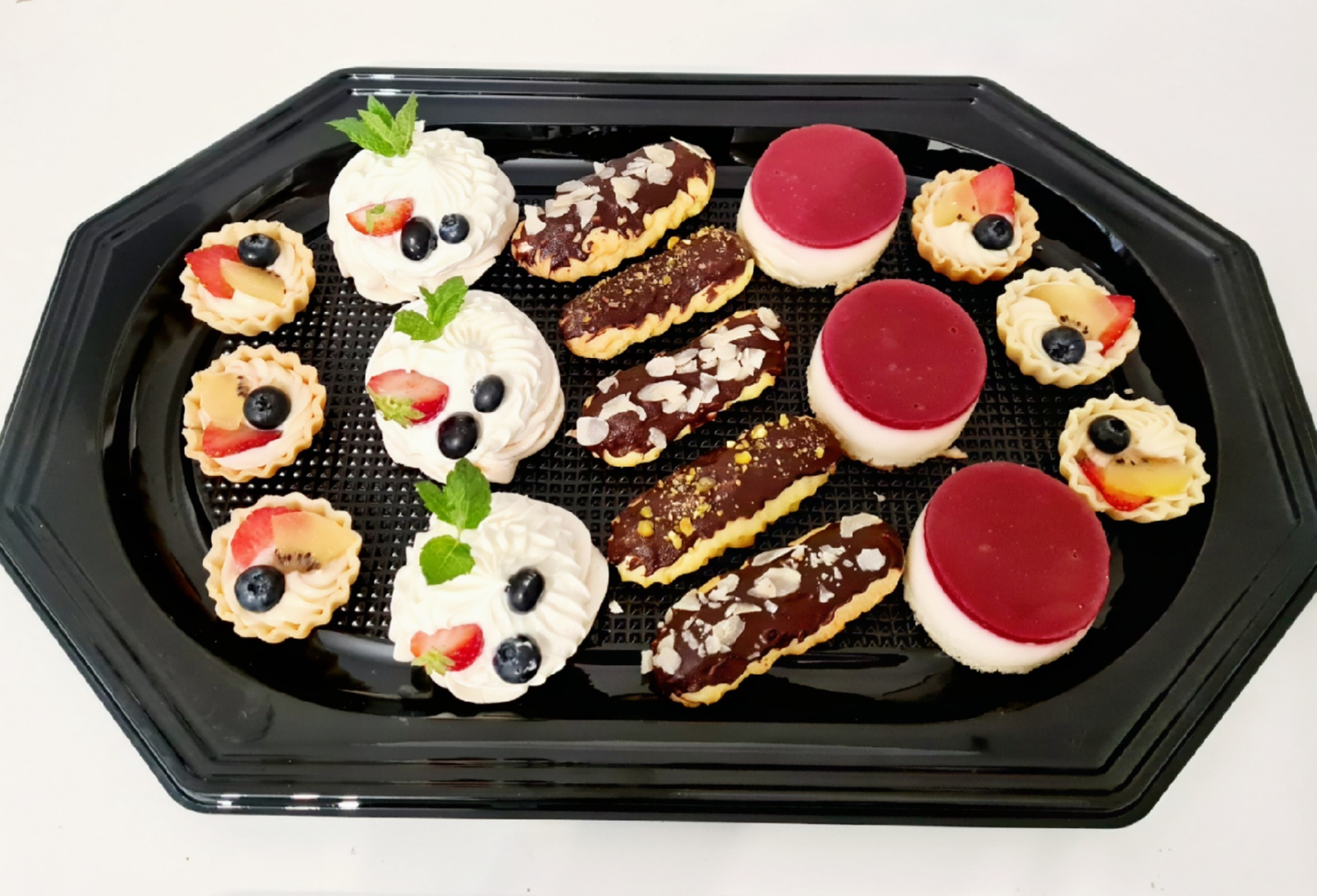 Elegancka czarna taca z różnorodnymi mini deserami: bezy z bitą śmietaną i owocami, eklerki z czekoladą i płatkami migdałów oraz mini tartaletki z kremem i owocami.