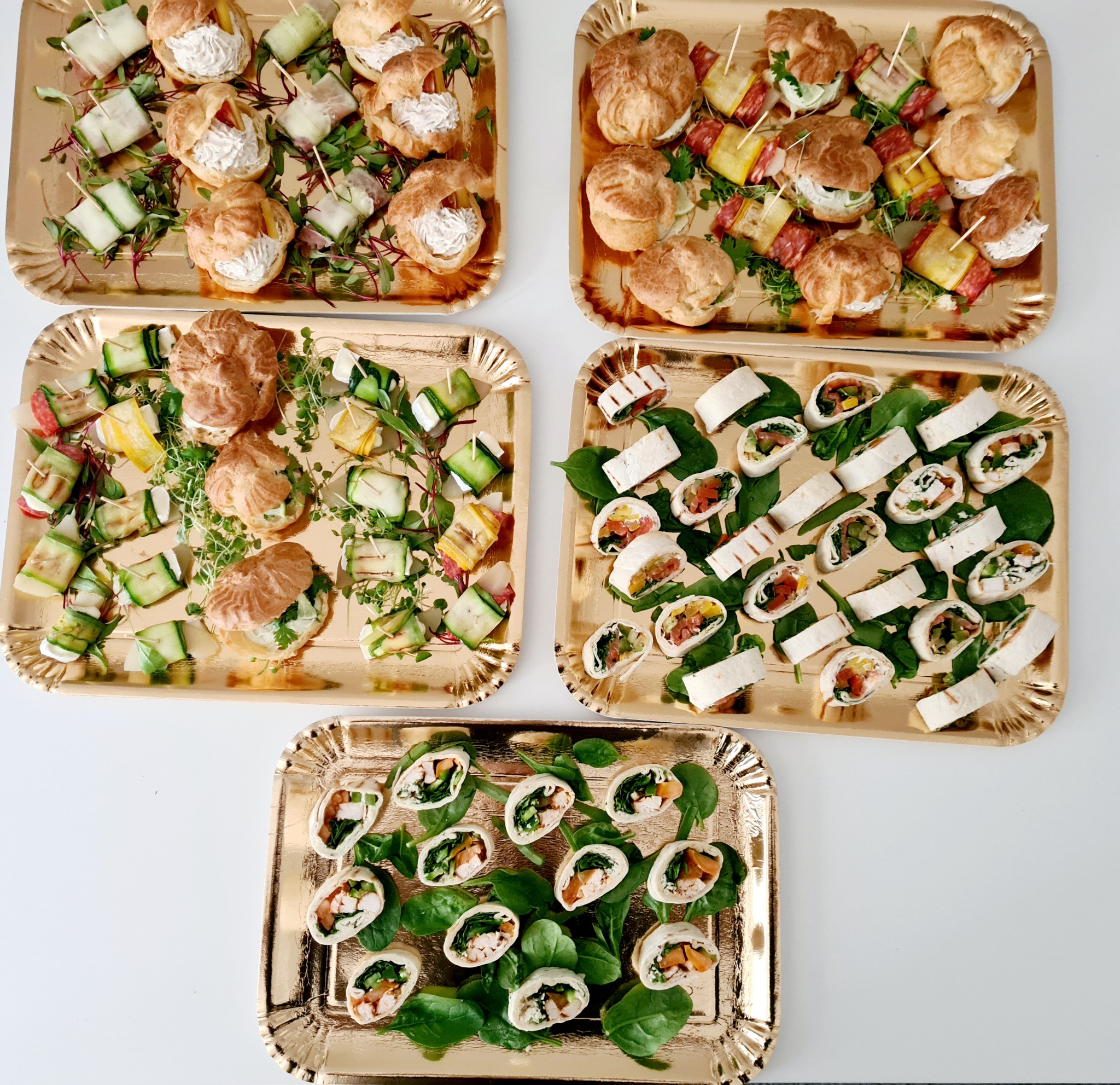 Zestaw pięciu złotych tac z różnorodnymi przekąskami: mini ptysie z kremowym farszem, roladki z warzywami i serem, koreczki z ogórka i szynki, całość udekorowana świeżymi ziołami.