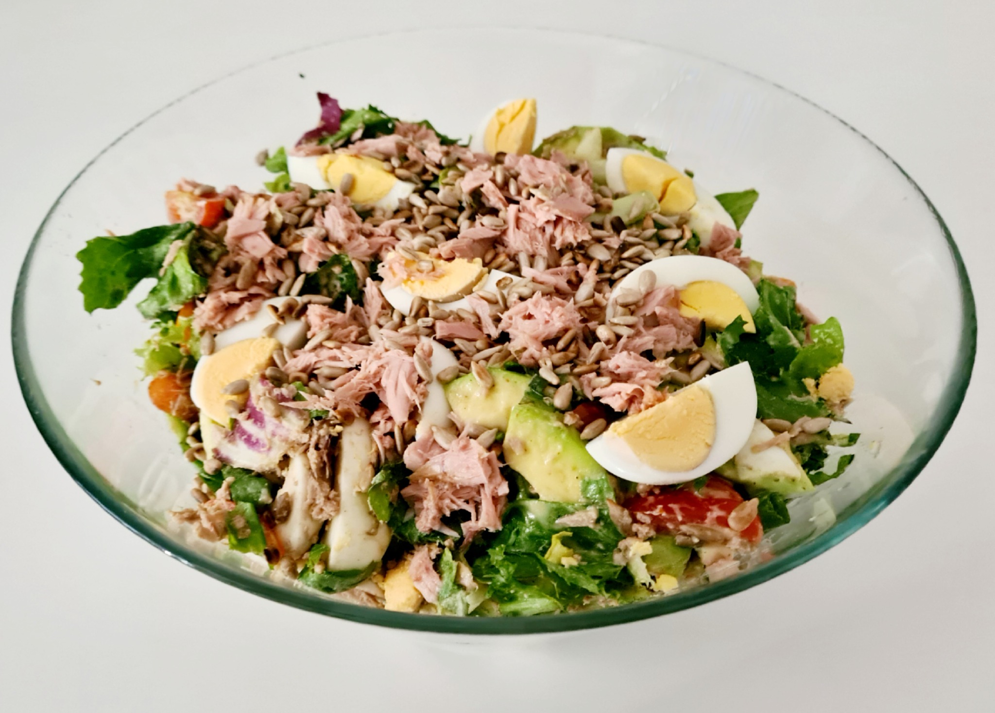Sałatka z tuńczykiem, jajkiem na twardo, awokado, pomidorami, sałatą i nasionami słonecznika, podana w szklanej misce na białym tle.