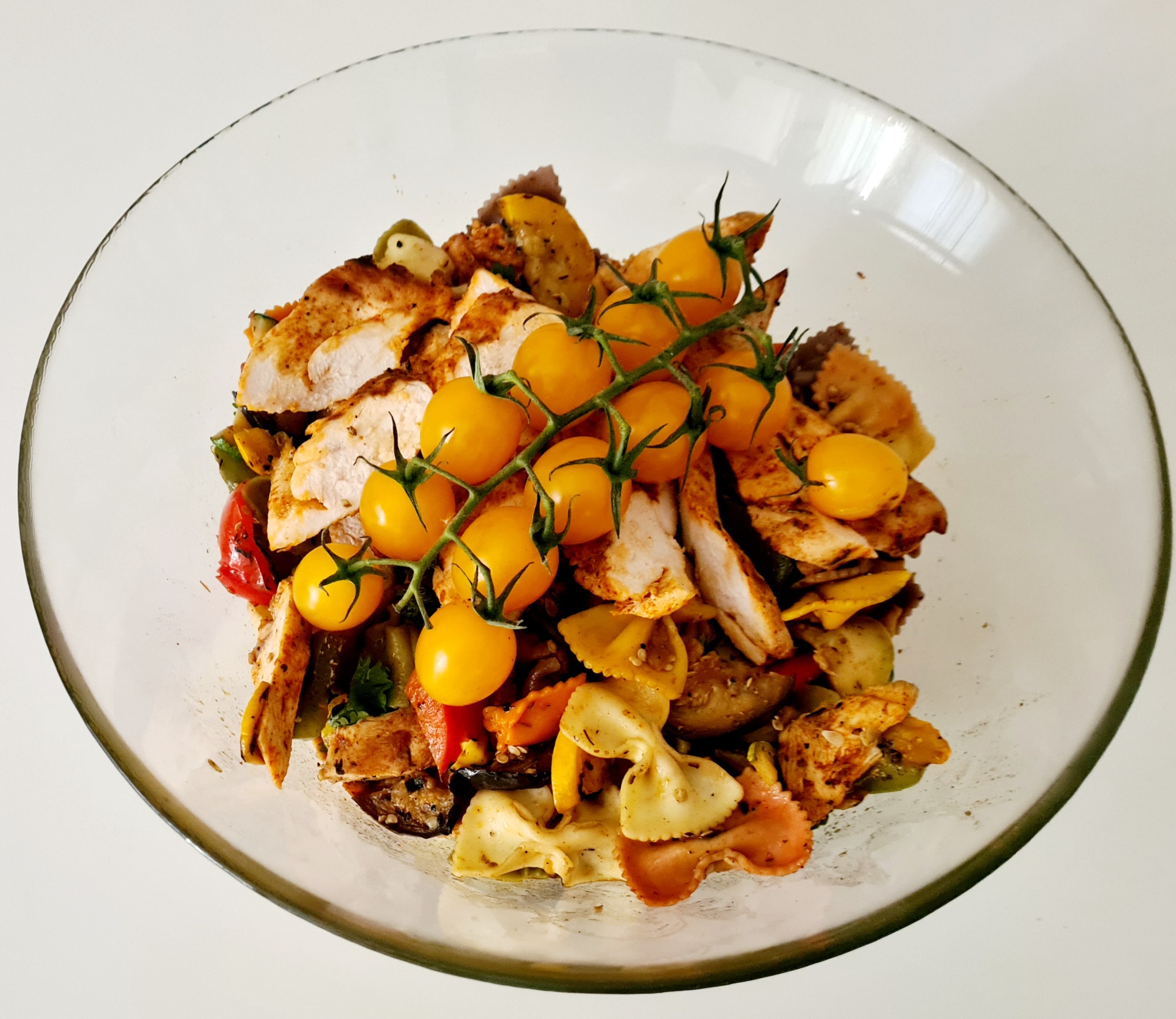 Sałatka z makaronem farfalle, grillowanym kurczakiem i pomidorkami koktajlowymi na gałązce, podana w szklanej misce.