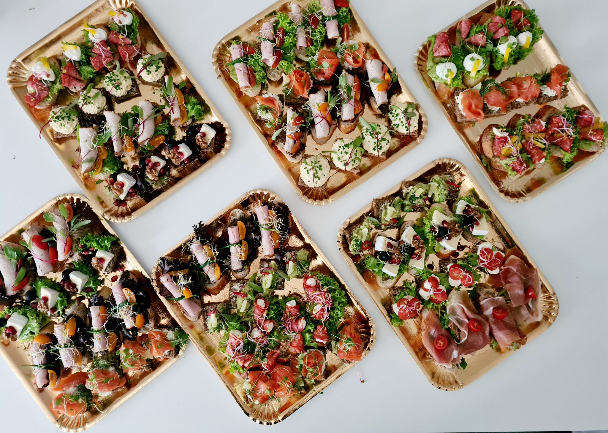 Różnorodne kanapki na sześciu złotych tacach, z wędlinami, serami, warzywami i owocami, ułożone na białym tle.