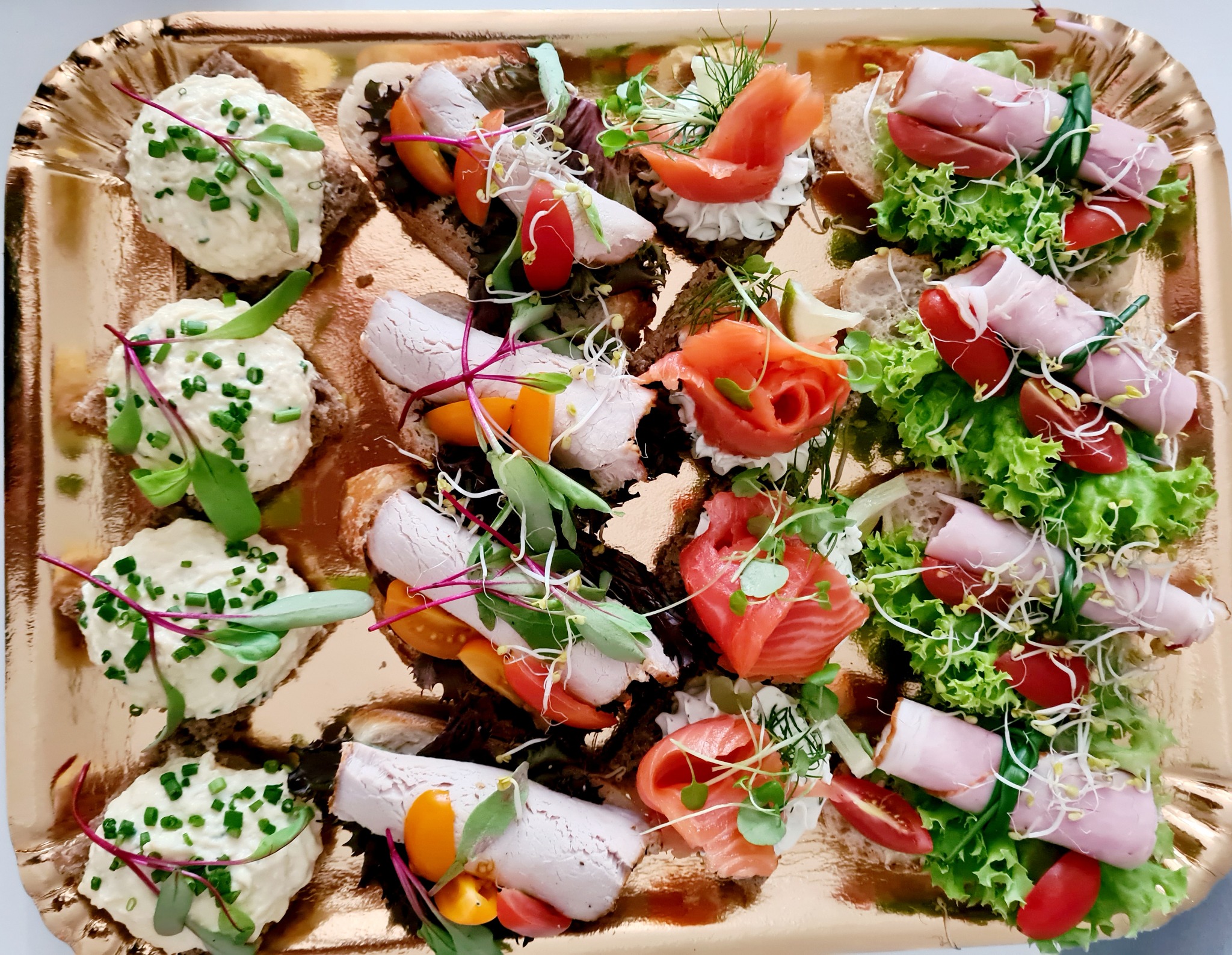 Elegancko podane, różnorodne kanapki cateringowe na złotej tacy, z dekoracjami z ziół i kiełków, w tym z pastą jajeczną, wędliną i łososiem.