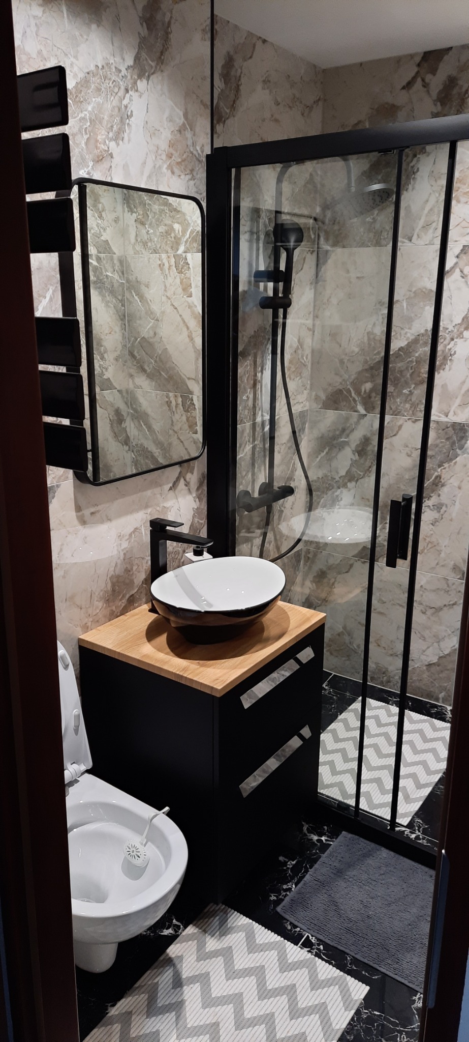 Nowoczesna łazienka z kabiną prysznicową typu walk-in, umywalką nablatową na szafce z blatem drewnopodobnym, grzejnikiem drabinkowym i toaletą. Na podłodze czarny marmur i dywanik w zygzaki.