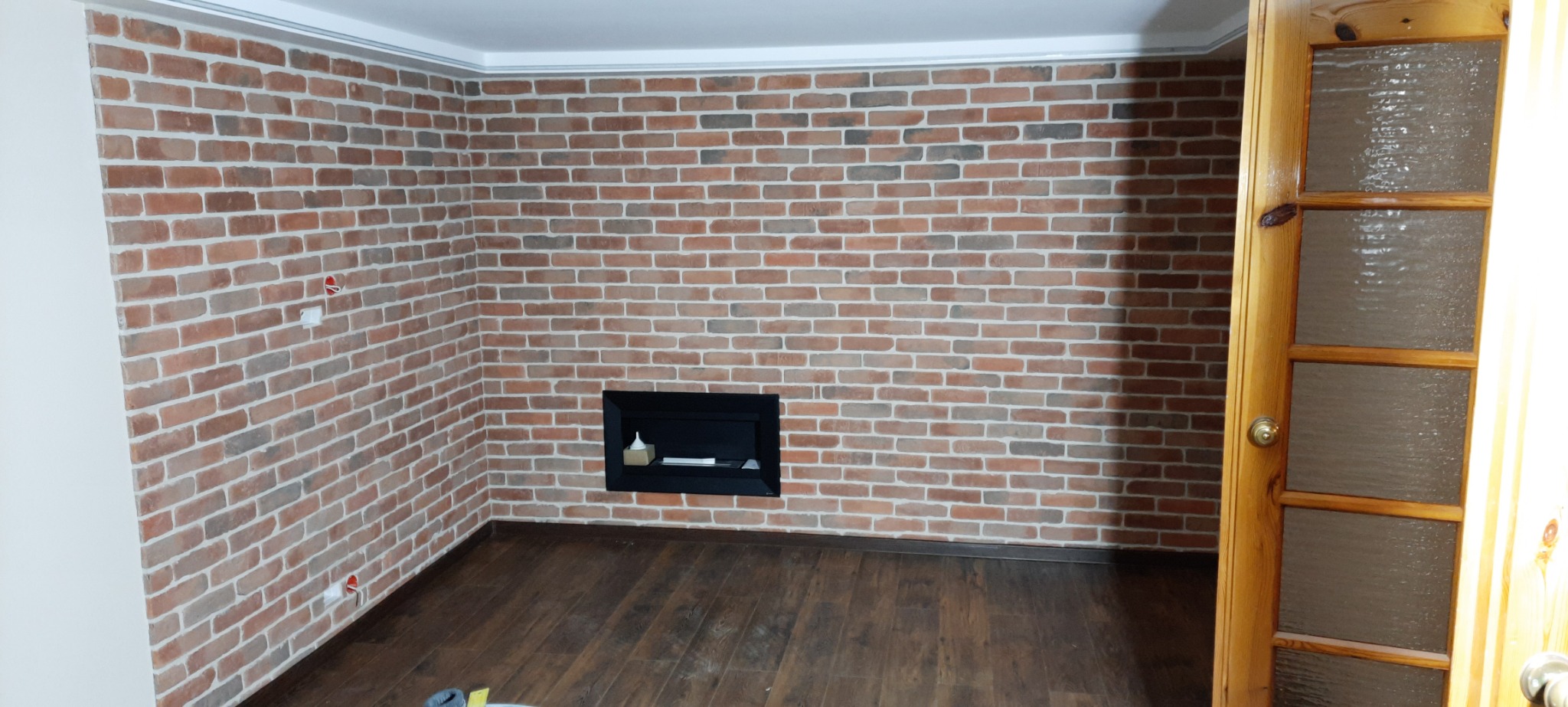 Ściana z cegły klinkierowej w odcieniach czerwieni i brązu, z wnęką na kominek, drewniana podłoga i częściowo widoczne drewniane drzwi z szybami, widoczne gniazdka elektryczne.