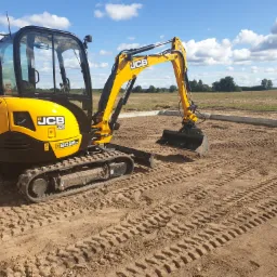 Żółta minikoparka JCB na ziemi z wyraźnymi śladami gąsienic, w tle widoczne pole i błękitne niebo z chmurami, obok ułożone betonowe obrzeża.