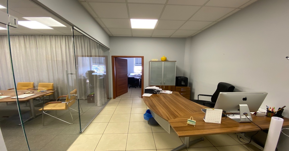 Wnętrze biura z przeszkloną salą konferencyjną po lewej stronie, biurkiem z komputerem iMac, kalendarzem i żółtą dekoracją na szafce w tle.